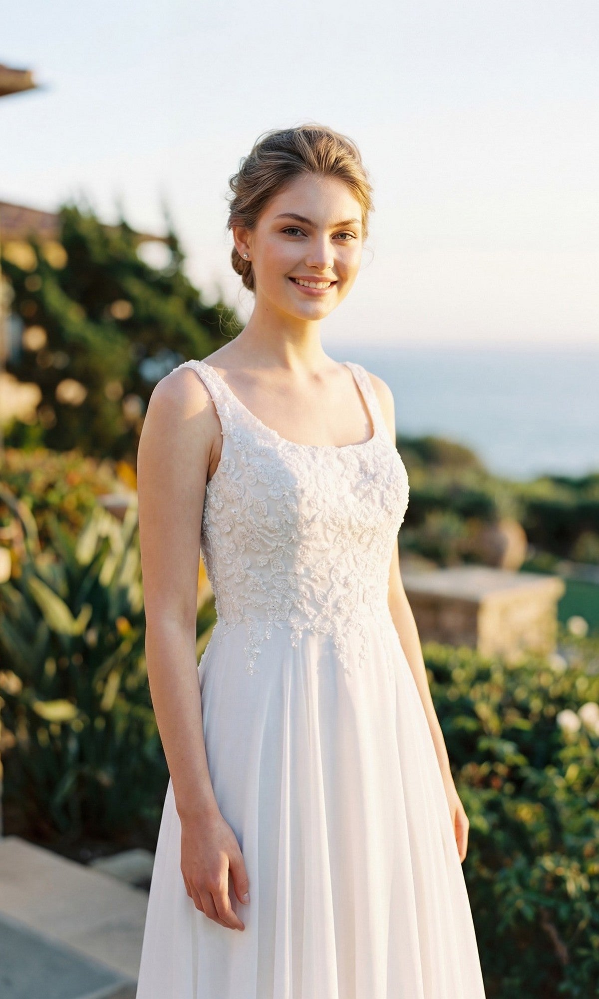 Wearing the Juliet JTM21LW long formal dress, a woman stands outdoors in a white, sleeveless, embroidered A-line gown, smiling with greenery and the ocean in the background.